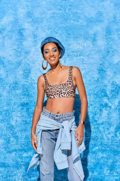 Усміхнена афро-американська модель з закритими очима і яскравим макіяжем, позує в береті, зверху з візерунком тварин і джинсами на синьому текстурованому фоні, стильний джинсовий одяг — стокове фото
