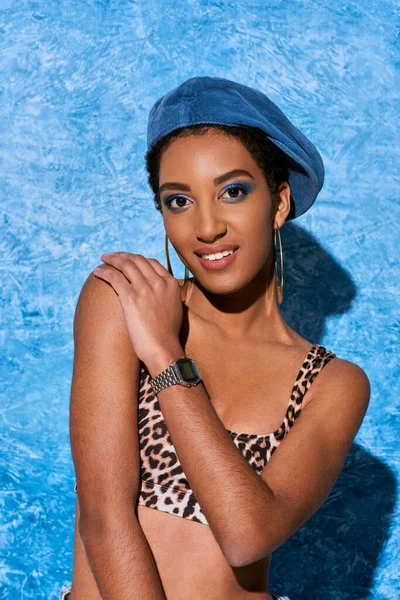 Portrait de femme afro-américaine souriante et à la mode en béret, boucles d'oreilles dorées et haut avec motif animal regardant la caméra sur fond texturé bleu, tenue en denim élégant — Photo de stock
