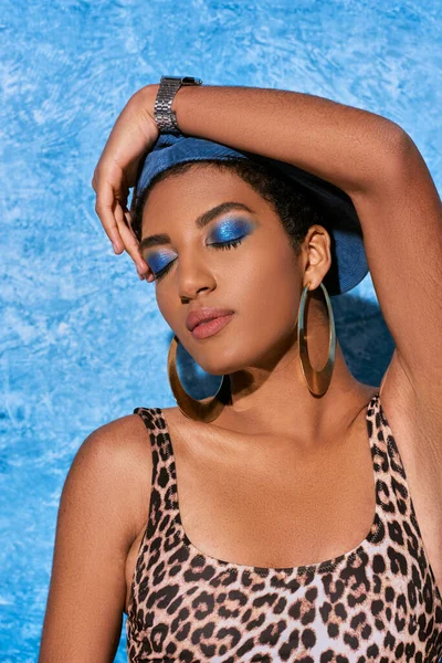 Portrait of trendy african american woman with bold makeup and golden earrings posing in top with animal print and touching beret on blue textured background, stylish denim attire — Stock Photo