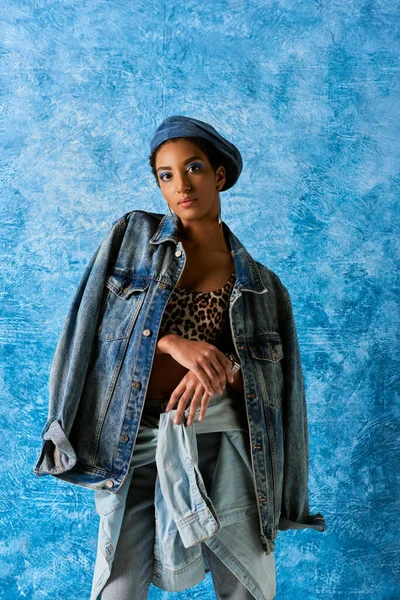 Giovane donna afroamericana in berretto guardando la fotocamera mentre posa in abiti in denim e top con motivo animale su sfondo blu testurizzato, elegante abbigliamento in denim — Foto stock