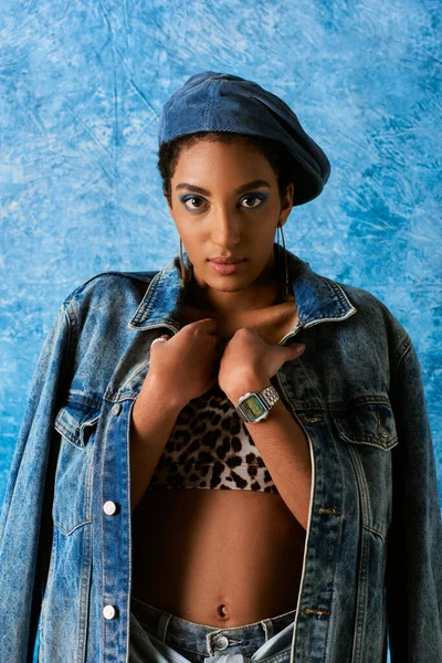 Porträt einer trendigen afrikanisch-amerikanischen Frau mit lebhaftem Make-up, die in Baskenmütze und Jeansjacke posiert, während sie in die Kamera blickt und auf blauem strukturiertem Hintergrund steht, stylische Jeans-Kleidung — Stockfoto