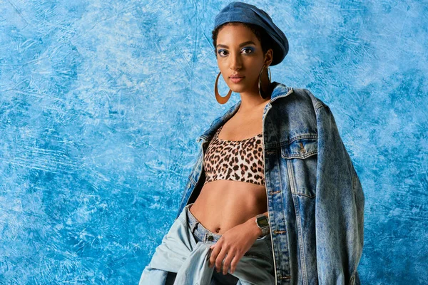 Сучасна афроамериканська модель в береті та золотих сережках з верхнім одягом з леопардовим принтом, джинсовою курткою та сорочкою на синьому текстурованому фоні, стильним джинсовим вбранням — стокове фото