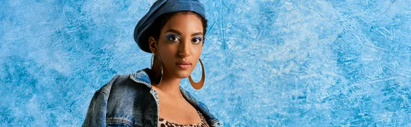 Moderna donna afroamericana in berretto e orecchini dorati in posa in giacca di denim e guardando la fotocamera su sfondo blu strutturato, elegante abbigliamento in denim, banner — Foto stock
