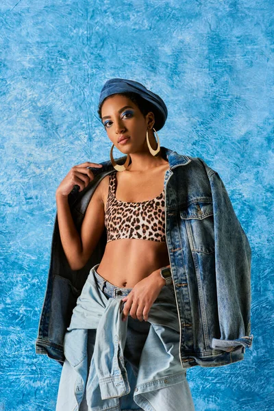 Selbstbewusste afrikanisch-amerikanische Frau mit Baskenmütze, goldenen Ohrringen und Top mit Leopardenmuster, die in die Kamera schaut und Jeansjacke auf blauem strukturiertem Hintergrund trägt, stylische Jeans-Kleidung — Stockfoto