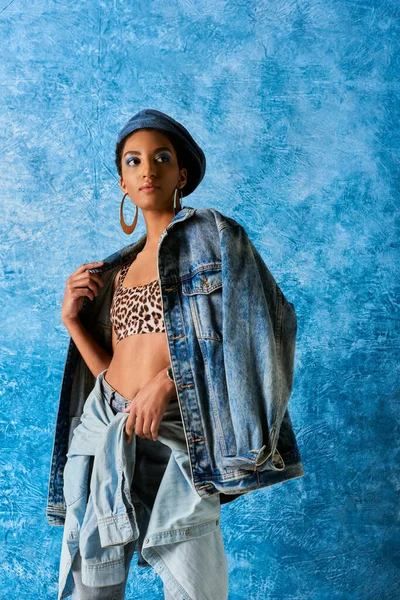 Modern african american woman with bold makeup and beret wearing denim jacket, top with animal print and jeans while standing on blue textured background, stylish jeans look — Stock Photo