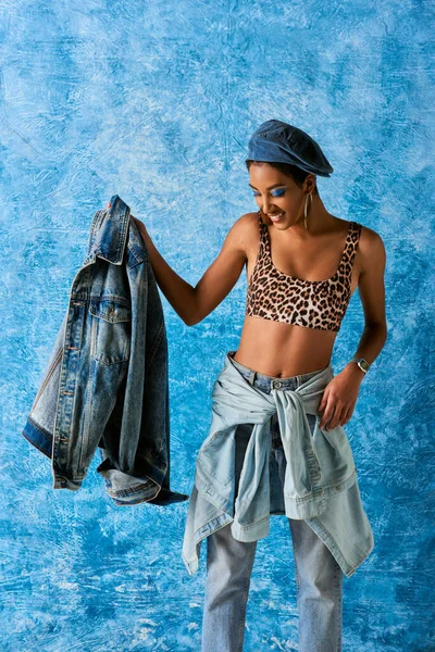 Sorrindo mulher afro-americana em boina, top com estampa de leopardo e jeans segurando jaqueta de ganga enquanto em pé sobre fundo azul texturizado, traje de ganga elegante — Fotografia de Stock