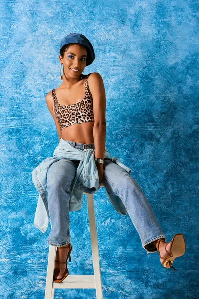 Full length of cheerful african american woman in beret, top with animal print and jeans sitting on chair and looking at camera on blue textured background, stylish denim attire — Stock Photo