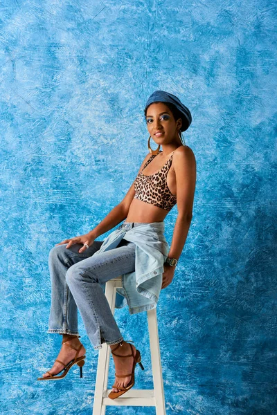 Positive african american woman with bold makeup and beret looking at camera while posing in top with animal print and jeans while sitting n chair on blue textured background, stylish denim attire — Stock Photo