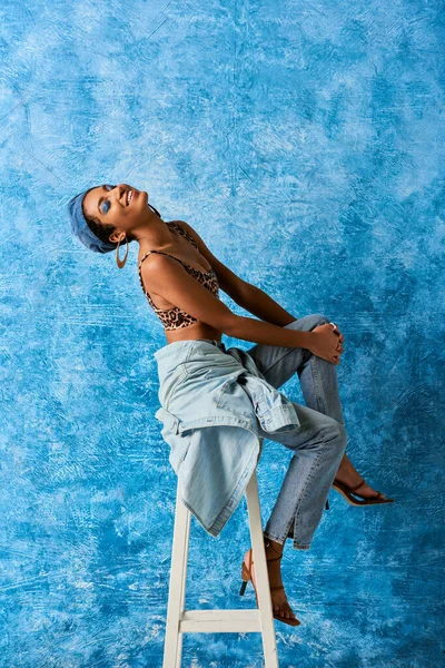 Positives afrikanisch-amerikanisches Model mit lebhaftem Make-up und goldenen Ohrringen, Oberteil mit Animal-Print und Jeans auf Stuhl sitzend auf blauem strukturiertem Hintergrund, stylische Jeans-Kleidung — Stockfoto