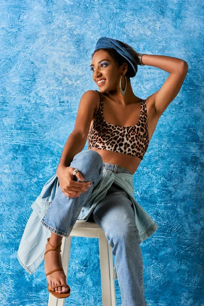 Trendy and positive african american woman in beret and golden earring wearing top with leopard print and jeans while sitting on chair on blue textured background, stylish denim attire — Stock Photo