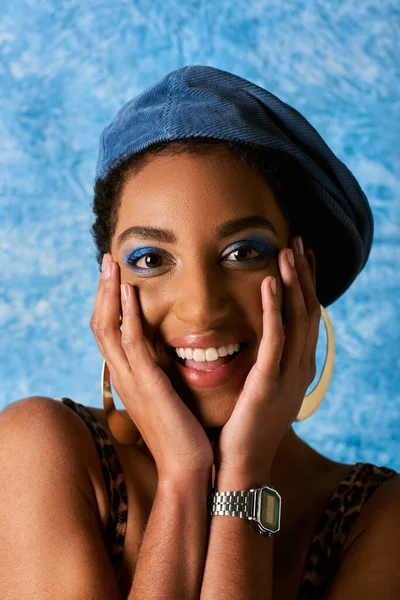 Portrait of positive african american woman with bold makeup, beret and golden earrings touching cheeks on blue textured background, stylish denim attire — Stock Photo