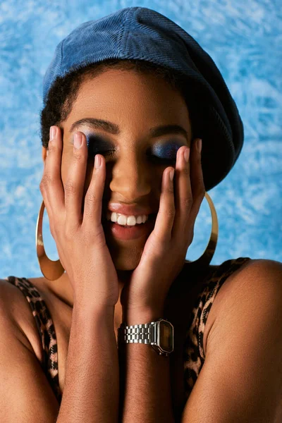 Porträt einer positiven afrikanisch-amerikanischen Frau mit lebhaftem Make-up und Baskenmütze, die Wangen berührt und die Augen schließt auf blauem, strukturiertem Hintergrund, stylische Jeans-Kleidung — Stockfoto