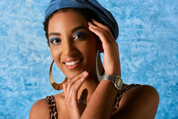 Modelo afroamericano de moda y sonriente con maquillaje audaz, pendientes de oro y boina posando y mirando a la cámara sobre fondo azul texturizado, elegante atuendo de mezclilla - foto de stock