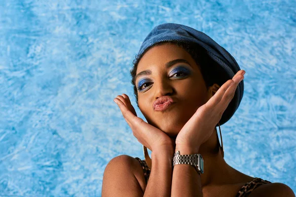 Portrait of trendy african american woman with bold makeup, golden earrings and beret pouting lips while standing on blue textured background, stylish denim attire — Stock Photo