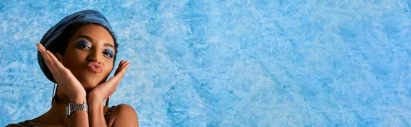 Modische afrikanisch-amerikanische Frau mit lebhaftem Make-up, goldenen Ohrringen und Barett schmollenden Lippen vor der Kamera auf blauem strukturiertem Hintergrund, stylische Jeans-Kleidung, Banner — Stockfoto