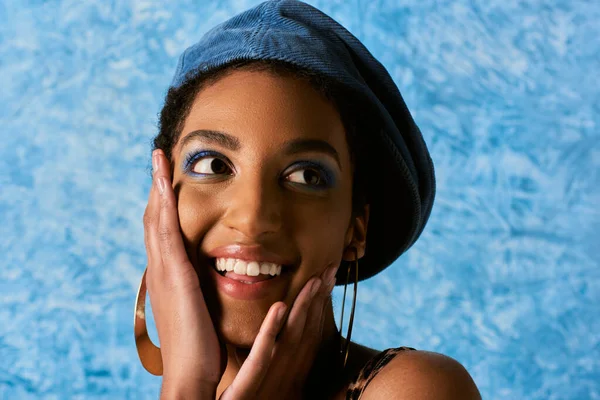Портрет позитивної афроамериканської моделі з яскравим макіяжем, беретом і сережками, які торкаються щоки і дивляться на синій текстурований фон, стильний джинсовий одяг — стокове фото