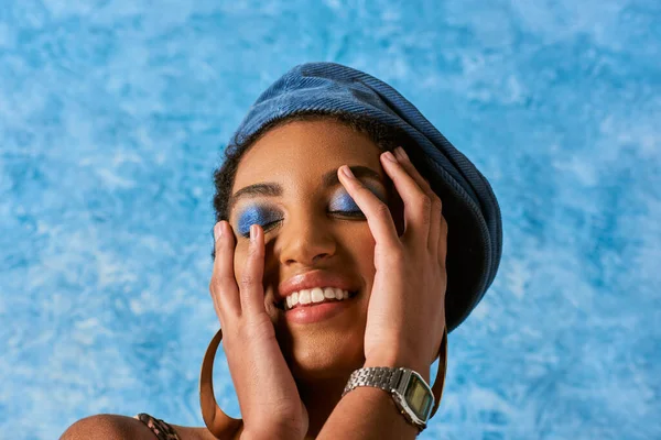 Femme afro-américaine gaie et élégante avec un maquillage vif portant béret en denim et boucles d'oreilles dorées tout en touchant le visage sur fond texturé bleu, tenue en denim élégant — Photo de stock