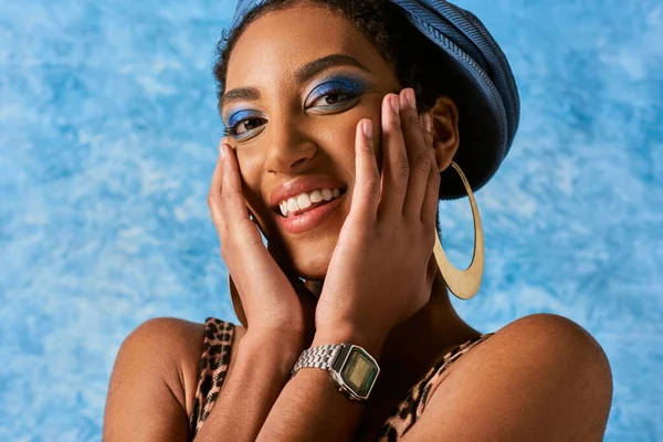 Retrato de modelo americano africano alegre com maquiagem vívida posando em brincos dourados e boina de ganga enquanto olha para a câmera em fundo azul texturizado, traje de ganga elegante — Fotografia de Stock