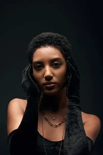 Portrait of fashionable and short haired african american woman with makeup and golden necklaces wearing gloves and dress while standing isolated on black, high fashion and evening look — Stock Photo