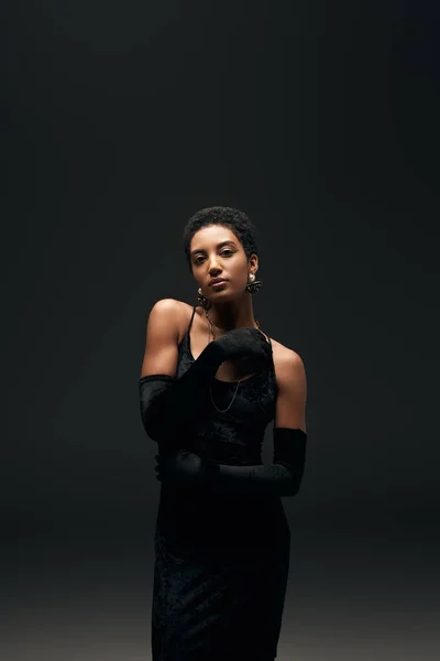 Elegante mujer afroamericana en guantes, collares dorados y pequeño vestido mirando a la cámara mientras está de pie aislado en negro, alta moda y look de noche - foto de stock