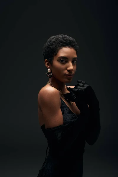 Elegant short haired african american woman with evening makeup posing in dress, gloves and golden accessories isolated on black, high fashion and evening look — Stock Photo
