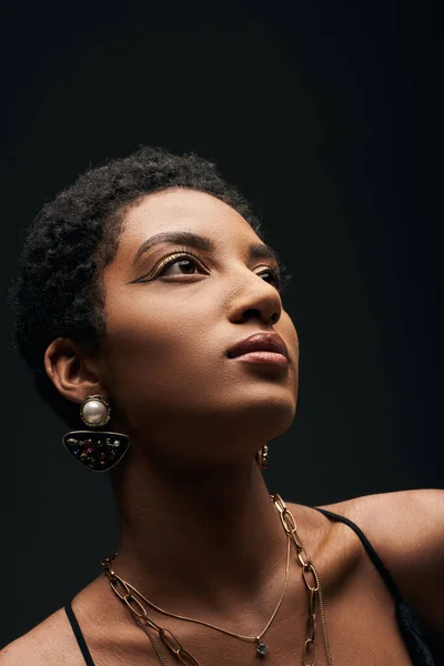 Retrato de mujer afroamericana elegante y de pelo corto con maquillaje de noche y accesorios dorados mirando hacia otro lado y de pie aislado en negro, alta moda y aspecto de noche - foto de stock