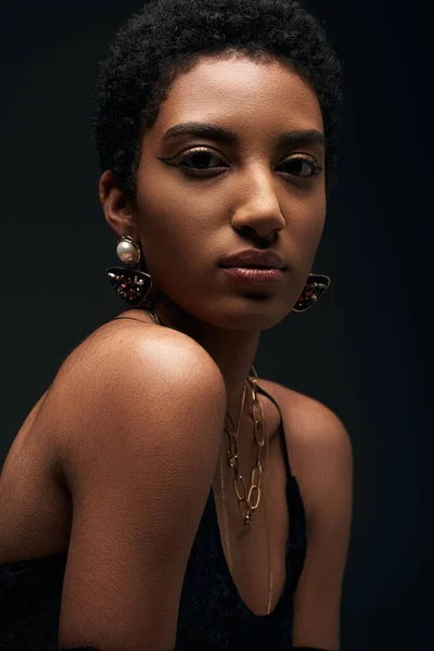Elegante modelo afroamericano joven en vestido de noche, maquillaje y accesorios dorados mirando a la cámara aislada en negro con iluminación, alta moda y aspecto de noche - foto de stock