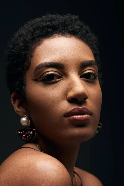 Vista de cerca de la mujer afroamericana joven y de pelo corto con pendientes y maquillaje de noche mirando a la cámara aislada en negro con iluminación, alta moda y aspecto de noche - foto de stock