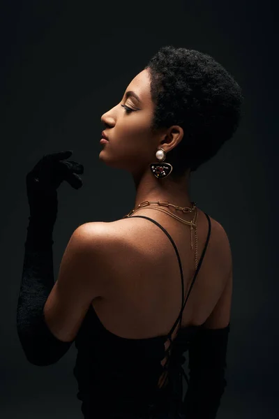 Вид збоку модна афроамериканська жінка з золотими аксесуарами та вечірнім платтям, що виглядає ізольовано на чорному з освітленням, високою модою та вечірнім виглядом — стокове фото
