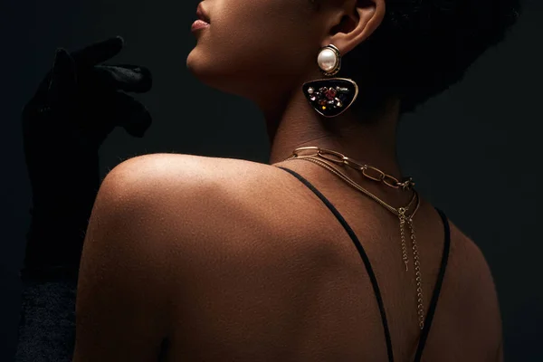 Vista recortada do elegante modelo afro-americano com acessórios dourados elegantes posando enquanto estava isolado em preto com iluminação, alta moda e olhar à noite — Fotografia de Stock