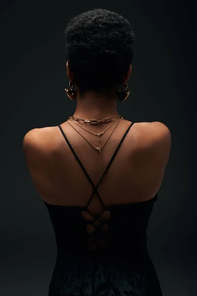 Back view of fashionable african american woman with short hair and golden accessories standing isolated on black with lighting, high fashion and evening look, jewelry, feminine, sensuality — Stock Photo