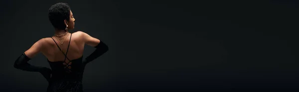 Vista lateral do moderno modelo da américa africana em luvas elegantes e acessórios dourados tocando quadris enquanto estava isolado em preto, alta moda e olhar à noite, banner — Fotografia de Stock