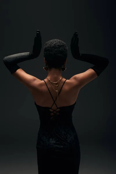 Visão traseira da mulher americana africana elegante em vestido de noite, luvas e acessórios dourados posando isolado no preto com iluminação, alta moda e olhar à noite, sensualidade feminina — Fotografia de Stock