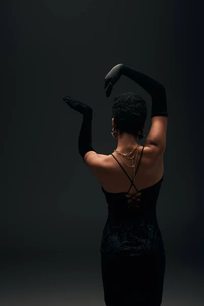 Vue arrière de l'élégante femme afro-américaine aux cheveux courts en gants, robe de soirée et accessoires dorés posant isolée sur look noir, haute couture et soirée — Photo de stock