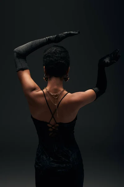 Back view of fashionable african american woman in gloves, golden accessories and evening dress posing while standing isolated on black, high fashion and evening look — Stock Photo