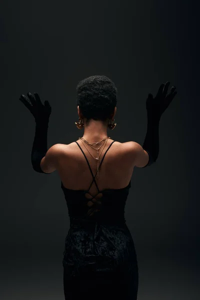 Vista posterior del modelo afroamericano de moda con accesorios dorados y ordenar el cabello posando en guantes y vestido de noche aislado en negro, alta moda y aspecto de noche - foto de stock
