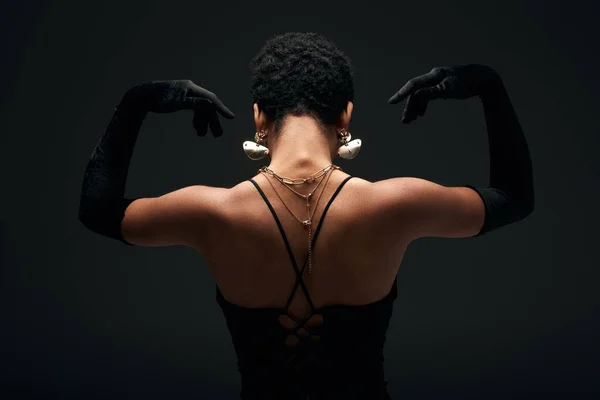 Vista posterior de la elegante mujer afroamericana en guantes y accesorios dorados posando en la luz mientras está de pie aislado en negro, alta moda y aspecto de noche - foto de stock