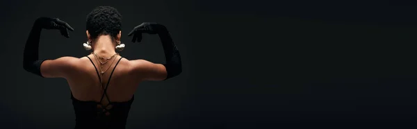 Rückansicht einer modischen jungen afrikanisch-amerikanischen Frau in Handschuhen und goldenen Accessoires, die bei Beleuchtung posiert, während sie isoliert auf schwarzem High Fashion und Abendlook steht, Banner — Stockfoto
