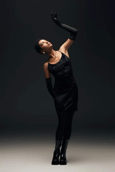Volle Länge der modischen jungen afrikanisch-amerikanischen Frau in elegantem Kleid und Handschuhen, die nach oben schaut, während sie auf schwarzem Hintergrund mit Beleuchtung, High Fashion und Abendlook posiert — Stockfoto