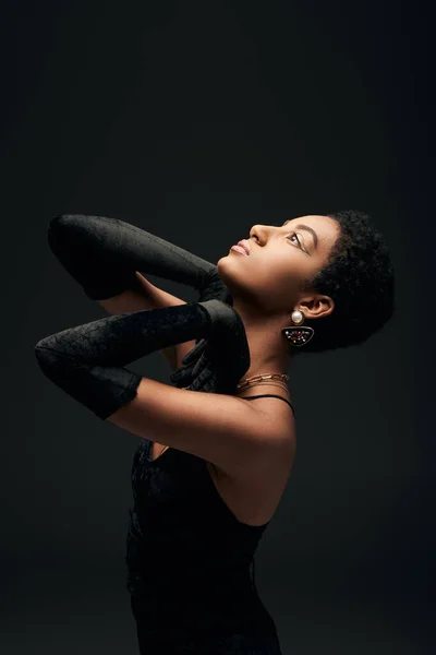 Вид збоку модної афроамериканської моделі в елегантних рукавичках і плаття, дивлячись вгору, стоячи під освітленням ізольовано на чорному, високій моді і вечірньому погляді — стокове фото