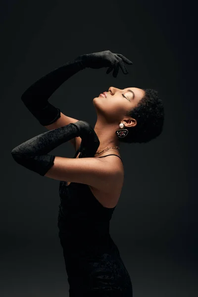 Вид збоку елегантна короткошерста афроамериканська жінка в рукавичках, сукнях і золотих аксесуарах, що позують із закритим оком ізольовано на чорному, високій моді і вечірньому погляді — стокове фото