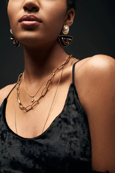 Cropped view of young and elegant african american model in dress and golden chains and earrings standing isolated on black under lighting, high fashion and evening look, details, close up — Stock Photo