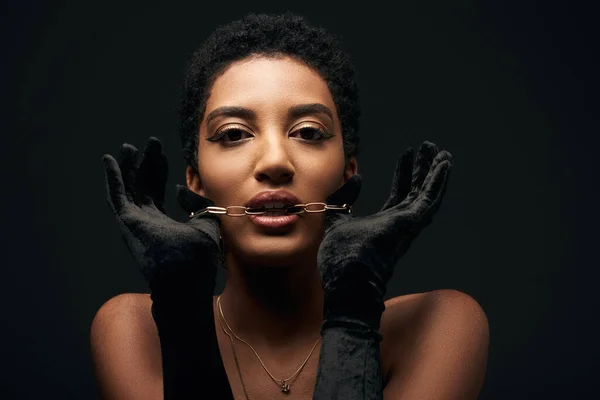 Fashionable and elegant african american model with evening makeup and gloves holding golden chain in mouth and standing isolated on black with lighting, high fashion and evening look — Stock Photo