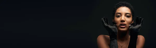 Modelo afroamericano de moda con maquillaje de noche que usa guantes y sostiene la cadena dorada cerca de la boca aislada en negro con iluminación, alta moda y apariencia nocturna, pancarta - foto de stock