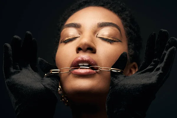 Portrait of fashionable african american woman with evening makeup wearing gloves and closing eyes while holding golden chain near mouth isolated on black, high fashion and evening look — Stock Photo