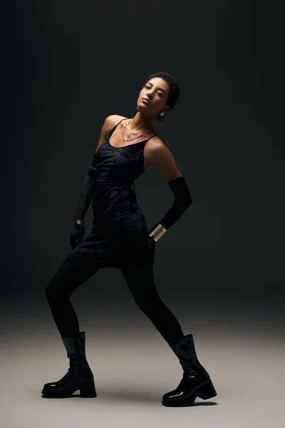 Longitud completa de la mujer afroamericana de moda en vestido pequeño, guantes y botas elegantes posando mientras mira a la cámara y de pie sobre fondo negro, alta moda y aspecto de noche - foto de stock