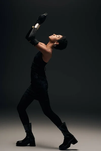 Vue latérale du modèle afro-américain à la mode en petite robe, gants et bottes posant tout en étant debout dans l'éclairage sur fond noir, look haute couture et soirée — Photo de stock