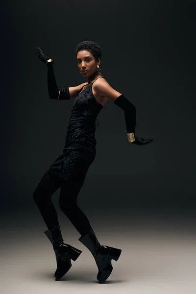 Selbstbewusstes junges afrikanisch-amerikanisches Model in kleinem Kleid, Handschuhen und Stiefeln posiert auf schwarzem Hintergrund mit Beleuchtung, High Fashion und Abendlook, Modemodel — Stockfoto