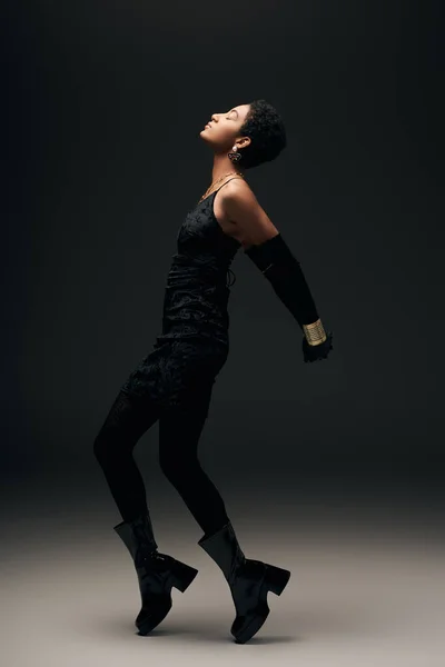 Seitenansicht einer modischen jungen Afroamerikanerin in Stiefeln, kleinem Kleid und goldenen Accessoires, die mit geschlossenen Augen auf schwarzem Hintergrund posiert, High Fashion und Abendlook — Stockfoto