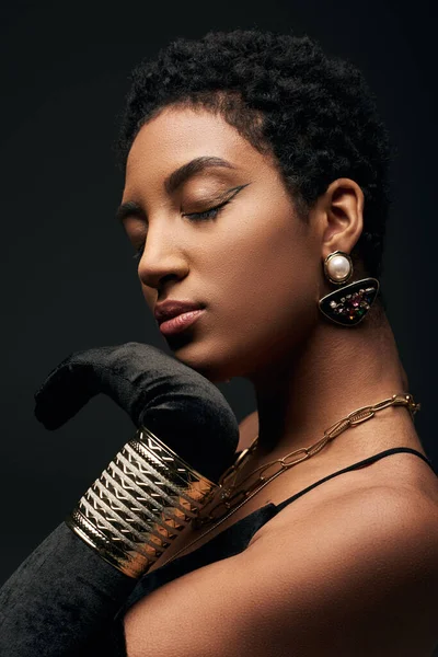 Portrait of fashionable african american woman in dress, glove and golden accessories touching chin while standing isolated on black, high fashion and evening look — Stock Photo